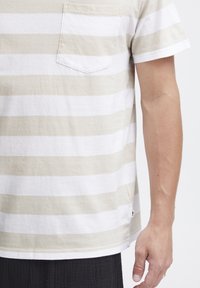 Camisa de rayas de manga corta en algodón beige y blanco. Presenta un bolsillo en el pecho, un corte relajado y aberturas laterales en el dobladillo.