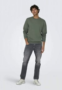 Jovem de pé, vestindo um moletom verde, jeans pretos desbotados e ténis brancos, contra um fundo claro e liso.