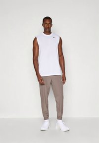 Sportiškas vyras stovi dėvėdamas baltą berankovę marškinėlę, šviesiai rudus bėgiojimo kelnes ir baltus sportinius batelius, priešais vienalytį šviesų foną.