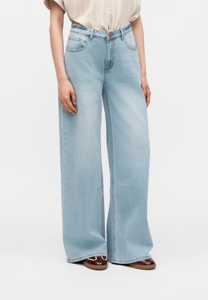 Persoon draagt lichtblauwe wijdvallende jeans met voorzakken en een gestreept T-shirt met korte mouwen, gecombineerd met bruine schoenen.