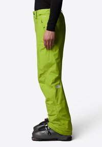 Limegröna skidbyxor med avslappnad passform, tillverkade av vattentåligt material. Utrustade med dragkedjefickor och förstärkta fållar. Kombineras med svarta skidstövlar.