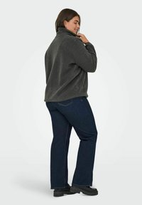 Kurtka polarowa w ciemnoszarym kolorze z wysokim kołnierzem, zestawiona z ciemnoniebieskimi rozszerzanymi dżinsami i czarnymi butami na platformie, widziana z boku.