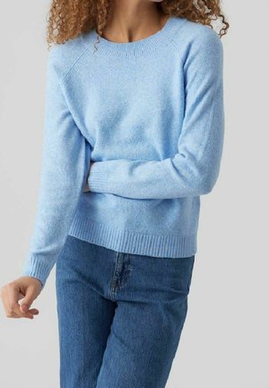 Vrouw met krullend haar, gekleed in een lichtblauwe trui met lange mouwen en blauwe spijkerbroek, poserend tegen een effen lichtgrijze achtergrond.