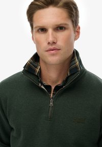 Forro polar verde con cuello de camisa de cuadros; cuenta con un logo en el pecho y una cremallera con un tirador, textura suave, diseño casual.