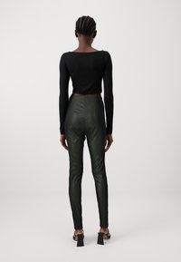 Un crop top noir à manches longues associé à un pantalon en simili cuir vert foncé taille haute, avec une texture lisse et une coupe ajustée. Des talons ouverts noirs.