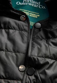 Közelkép egy fekete, steppelt dzsekiről, ezüst és fekete kapcsos gombokkal, valamint egy zöld címkével, amelyen a "Portland Outerwear Co." felirat olvasható.