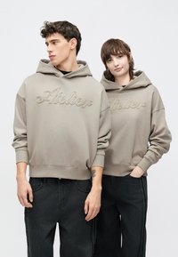 Túlméretezett bézs kapucnis pulóverek hímzett "Atelier" felirattal, fekete farmernadrággal párosítva, bordázott mandzsettákkal és laza szabással.