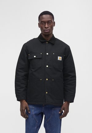 Homme portant une veste noire boutonnée avec quatre poches avant et un jean bleu, debout devant un fond clair uni.