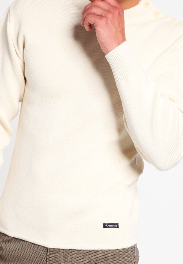 Sweter w kremowym kolorze z prążkowymi mankietami, zapięciem na trzy guziki przy szyi oraz naszywką z logo na dolnej lewej stronie. Miękka, dzianinowa tekstura.