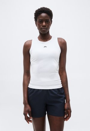 Ženska s kratkimi kodrastimi lasmi, oblečena v oprijeto belo brezrokavnik in mornarsko modre kratke hlače, stoji pred enobarvnim svetlim ozadjem.