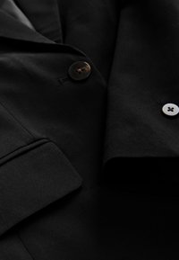 Černé sako s hladkou texturou, klopy s výstřihy a dvěma kontrastními knoflíky – jedním v tmavém rohovém provedení a jedním bílým.