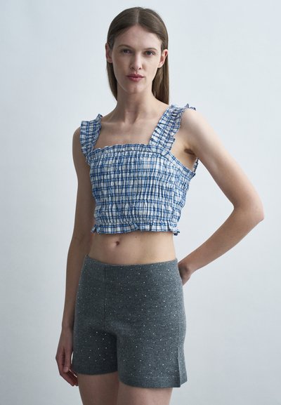 Femme portant un crop top à volants à carreaux bleus et blancs et un short en tricot gris avec de petits points blancs, debout devant un fond clair uni.