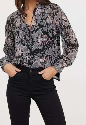 Femme portant un chemisier noir transparent avec des motifs floraux roses et gris et un pantalon noir taille haute, les mains dans les poches.