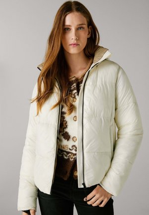 Giacca imbottita color crema con colletto alto, chiusura a zip e accenti scuri, indossata sopra un maglione in maglia marrone a fantasia.