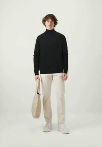 Un pull à col roulé noir, un pantalon crème et des baskets blanches, tenant un sac fourre-tout beige matelassé, présenté contre un fond neutre.