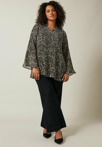Haut en imprimé léopard transparent noir et beige avec des manches évasées, associé à un pantalon large noir et des chaussures à talons hauts noires.