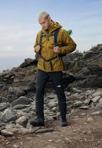 Olivovo zelená nepremokavá bunda s nastaviteľnými manžetami, čierne nohavice a pevnú čiernu turistickú obuv. Model stojí na kamenistom teréne.