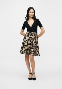 Μαύρο φόρεμα με εφαρμοστό μπούστο και φουσκωτή φούστα, με χρυσό λουλουδάτο σχέδιο. Κοντά μανίκια και βαθύ ντεκολτέ V. Μαύρα παπούτσια.