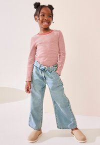 Röd och vit randig långärmad skjorta, ljusblå vida jeans med bälte, beige slip-on skor, mot en neutral bakgrund.