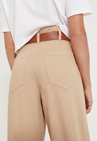 Pantalons beiges taille haute avec deux poches arrière et une ceinture en cuir marron. Le tissu semble doux avec une finition légèrement texturée.