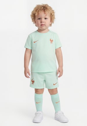 Tout-petit aux cheveux blonds bouclés portant un ensemble de football vert menthe avec des logos de coq orange, des chaussettes vert menthe et des baskets blanches, debout sur un fond blanc.