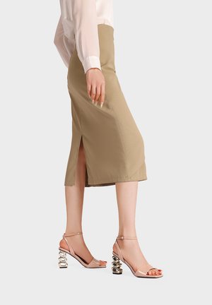 Donna che indossa una gonna midi beige con spacco laterale e sandali con tacco alto nudi con cinturini e tacchi metallici scolpiti, in piedi di profilo.