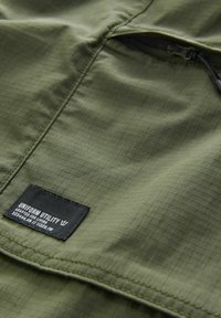 Olivovo zelená tkanina s textúrou mriežky, s vreckom na zips a čiernou nálepkou s nápisom "UNIFORM UTILITY ADAPTED FOR LIVING."