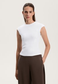 Biely rebrovaný top s krátkym rukávom a priliehavým strihom, s okrúhlym výstrihom. V kombinácii s hnedými nohavicami s vysokým pásom, ruka v vrecku.