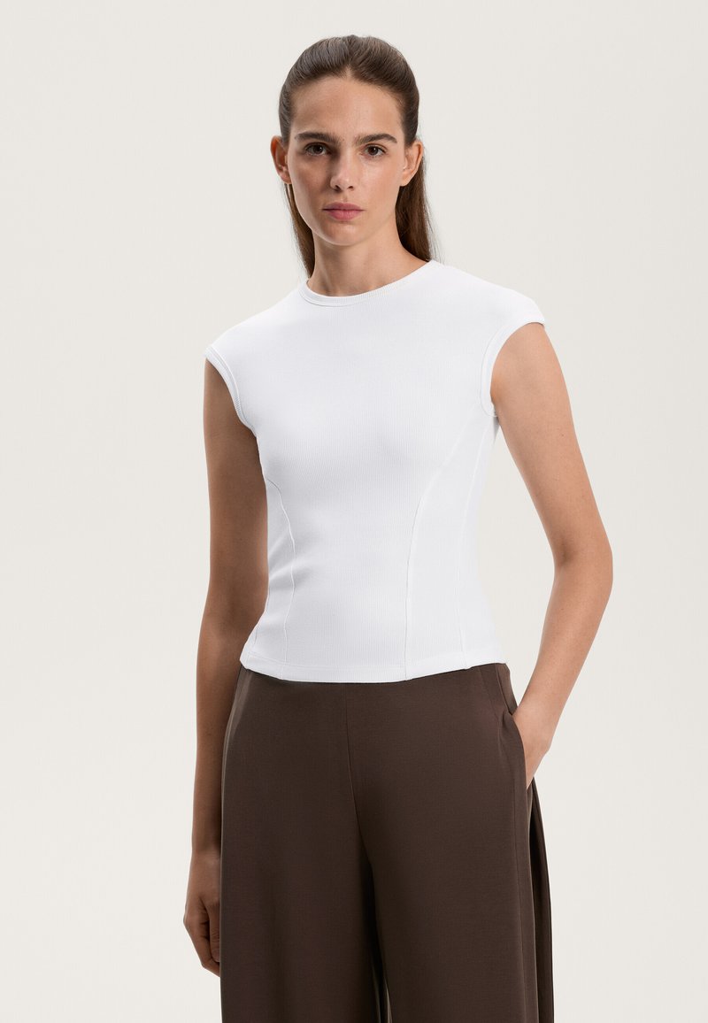 Biely rebrovaný top s krátkym rukávom a priliehavým strihom, s okrúhlym výstrihom. V kombinácii s hnedými nohavicami s vysokým pásom, ruka v vrecku.