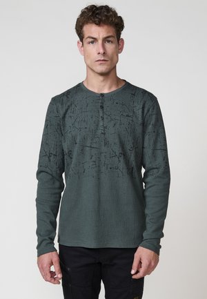 Camisa de manga larga en verde oscuro, con un patrón texturizado y acentos negros, y un cuello redondo con tapeta de tres botones.