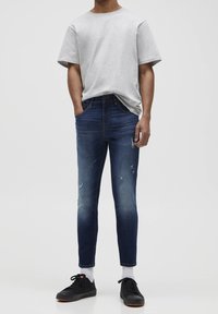 Camiseta de cuello redondo gris y vaqueros azul oscuro con detalles desgastados. Zapatillas negras, calcetines blancos, de corte ajustado y textura suave.