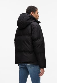 Sort pufferjakke med hætte, der har et quiltet design og ribbede manchetter, båret over blå jeans. Glat tekstur og kraftig isolering.