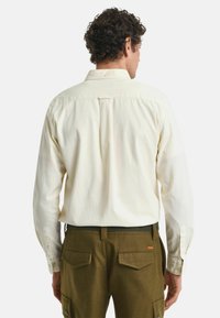 Cremfarbener Langarmhemd mit Button-Down-Kragen, Rückenfalten und Manschetten, getragen mit olivgrünen Cargo-Hosen mit Gesäßtaschen.