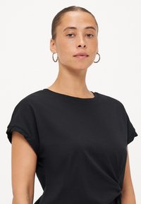 Zwarte T-shirt met opgerolde mouwen, gemaakt van zachte stof, met een ontspannen pasvorm en een knoopdetail aan de voorkant. Effen, geen patronen.
