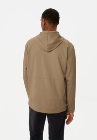 Brauner, leichter Hoodie mit strukturiertem Finish, ausgestattet mit einer Kapuze mit Kordelzug und Seitentaschen, getragen mit schwarzen Hosen, von hinten betrachtet.