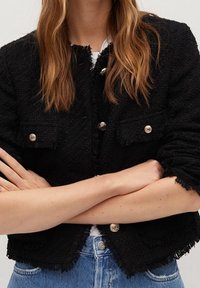 Musta tekstitetty takki, jossa on hopeanväriset nappi ja kaksi etutaskua, frayed reunat ja lyhyt muotoilu. Päällä valkoinen paita.