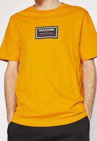 Muž v jasně oranžovém tričku s logem "Hilfiger American Classic Est. 1985 New York" na hrudi, zasunutém do černých kalhot.