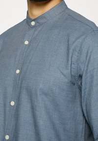 Camicia blu con colletto alla cinese, caratterizzata da un tessuto texturizzato e bottoni di colore chiaro. Vista ravvicinata che si concentra sulla parte superiore.
