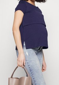 Marinblå kortärmad gravidblus med lagerdesign och sidoöppningar, tillsammans med ljusblå slitna jeans och en taupe handväska.