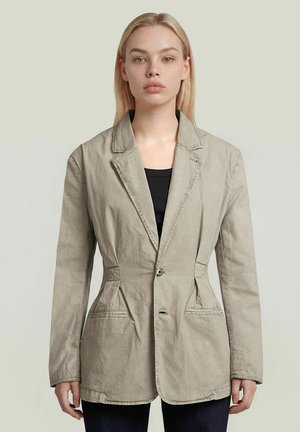 Jonge vrouw met stijl blond haar, die een beige blazer met één knoop draagt over een zwarte top, staand tegen een effen lichte achtergrond.