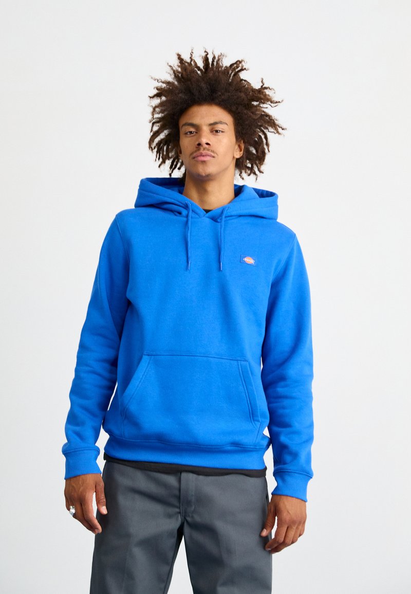 Sudadera de algodón azul con un bolsillo tipo canguro, capucha con cordones, puños acanalados y parche de logo en el pecho, combinada con pantalones grises.