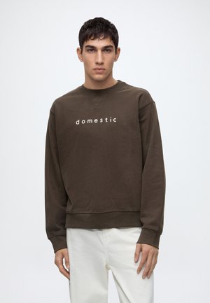 Jeune homme portant un sweat-shirt marron avec le mot "domestic" imprimé dessus et un pantalon blanc, debout devant un fond clair uni.
