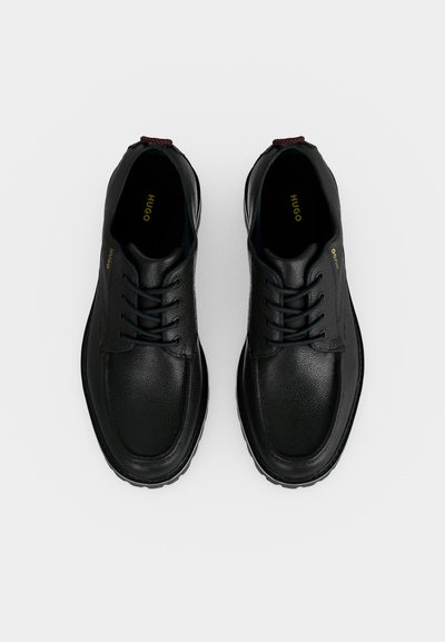 Paire de chaussures en cuir noir à lacets avec surface texturée et marquage "HUGO" à l’intérieur et sur les côtés, présentée vue de dessus sur fond blanc.