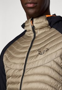 Veste doudoune beige avec un design matelassé, manches noires et une touche d'orange. Dispose d'une fermeture éclair à l'avant et d'un petit logo sur la poitrine.