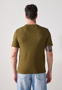 Tricou texturat de culoare verde măsliniu, cu mâneci scurte și guler rotund. Materialul are un aspect ribat, asociat cu blugi de culoare albastru deschis.