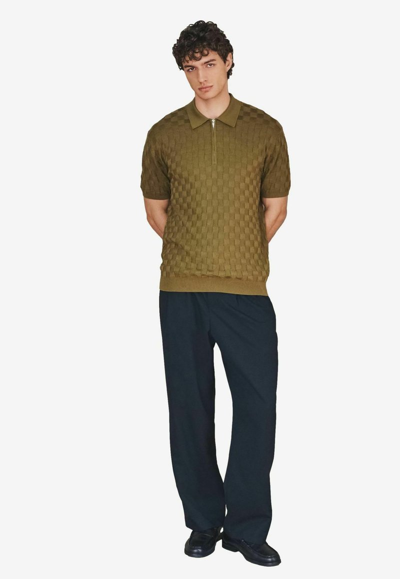 Polo manches courtes en tricot vert olive avec une fermeture éclair à l'avant et un motif damier texturé, associé à un pantalon noir ample et des chaussures noires.