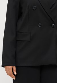 Μαύρο διπλό γιλέκο με δύο κουμπιά, δομημένη εφαρμογή και πλαϊνή τσέπη. Το ύφασμα φαίνεται λείο με ελαφριά γυαλάδα.