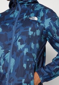 Veste coupe-vent bleue avec un motif abstrait texturé. Elle dispose d'une fermeture éclair, de poignets élastiques, et du logo The North Face sur la poitrine.