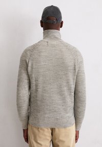 Grijze, ribbelige gebreide trui met een hoge kraag en strakke mouwen; aangevuld met een grijze pet. G gedragen met lichtgekleurde corduroy broek.