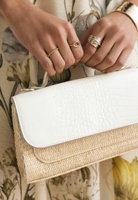 Borsa bianca con una pattina in pelle ecologica effetto coccodrillo testurizzata e base in paglia intrecciata. Presenta una maniglia superiore, fissata con dettagli in metallo dorato.